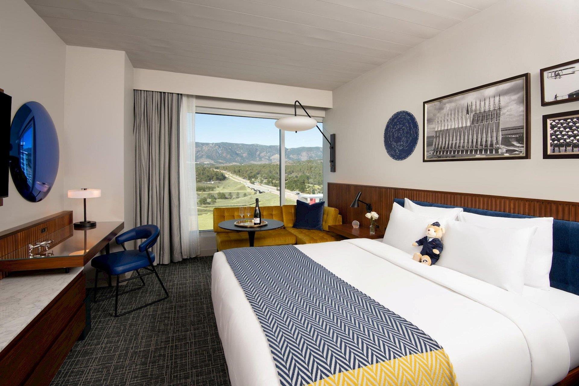 Mountain-view king room with white bedding, blue headboard, blue-and-yellow patterned throw, wood wall paneling, mustard window sofa, and desk area.