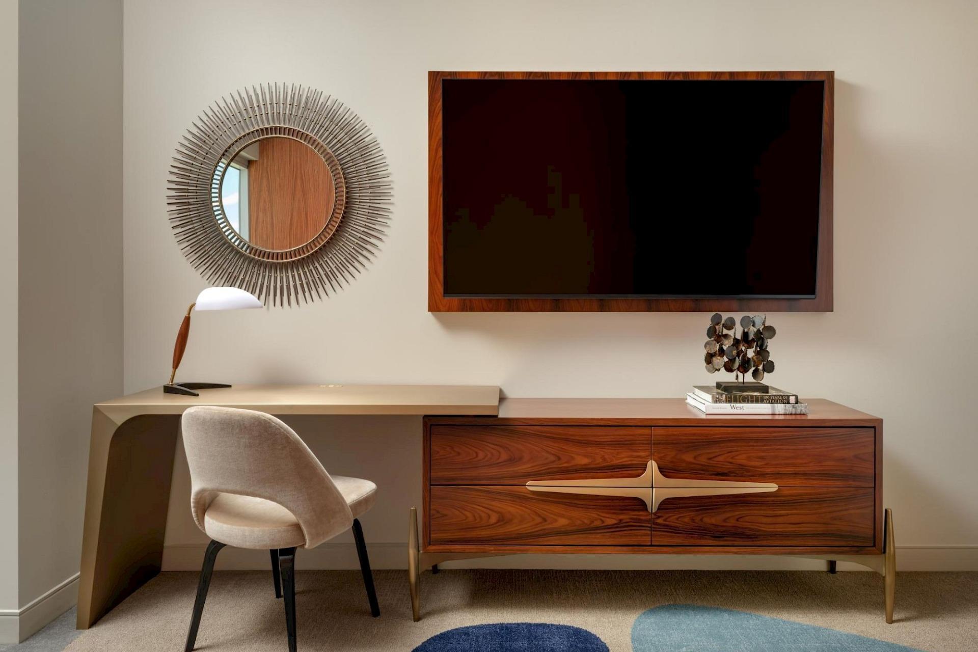 Guest room work area with a curved desk, upholstered chair, sunburst mirror, wall-mounted TV, and mid-century wood dresser with brass detailing.