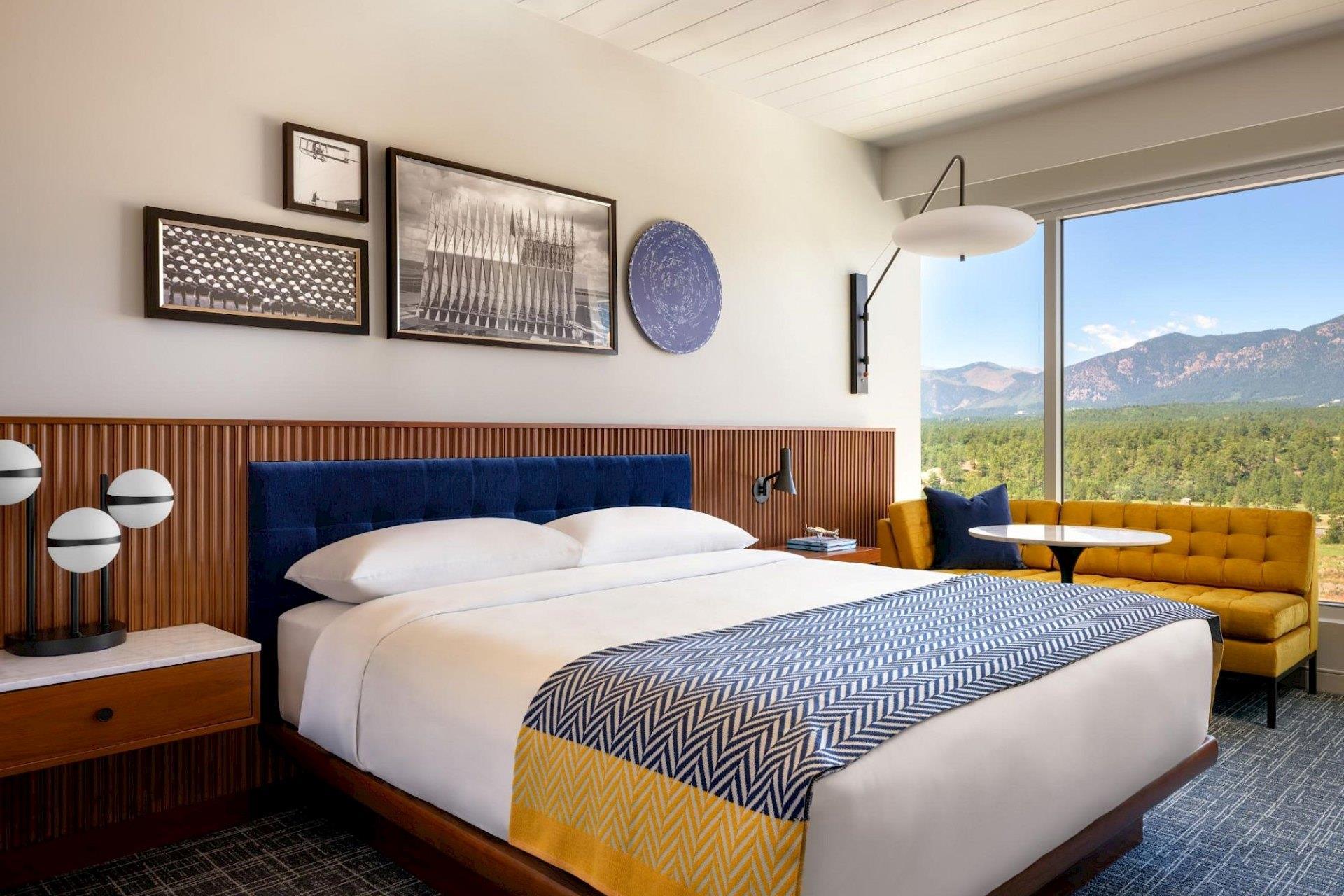 King guest room with white bedding, blue-and-yellow patterned throw, blue headboard, warm wood paneling, mustard sofa, and expansive mountain view.