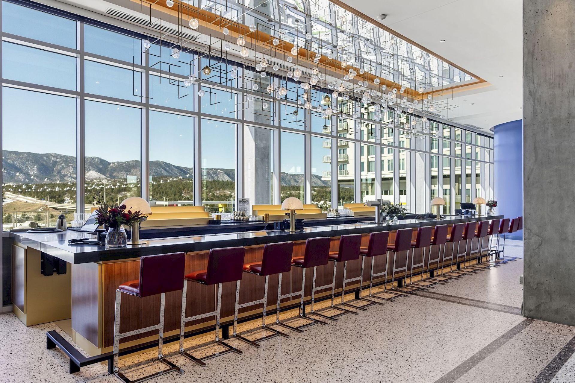 Long contemporary bar with burgundy stools, wood and brass finishes, suspended glass lighting, and floor-to-ceiling windows overlooking the mountains.