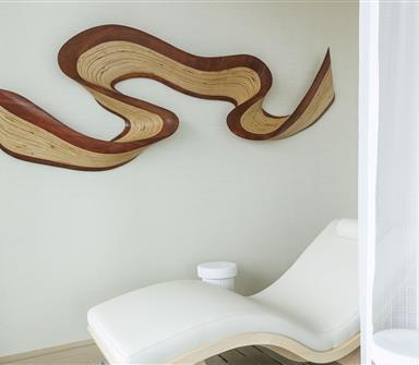 Spa relaxation area with a white contoured lounge chair beneath an abstract layered wood wall sculpture and sheer privacy curtain.