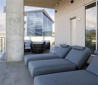 Outdoor terrace with cushioned gray lounge chairs, glass railings, and views of a modern glass building and distant mountains.