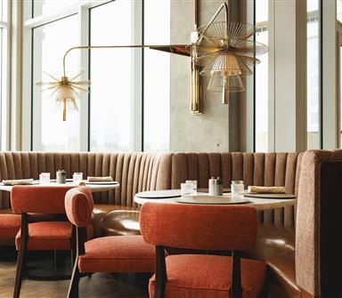Restaurant seating area with curved brown banquettes, coral upholstered chairs, round tables, sculptural brass wall lights, and floor-to-ceiling windows.