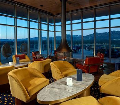 Upscale lounge with mustard-yellow chairs, red leather seating, marble tables, a central fireplace, and floor-to-ceiling windows overlooking mountains at dusk.