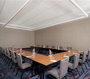 Modern meeting room with wood tables arranged in a U-shape, gray upholstered chairs, water service, and geometric blue patterned carpet.
