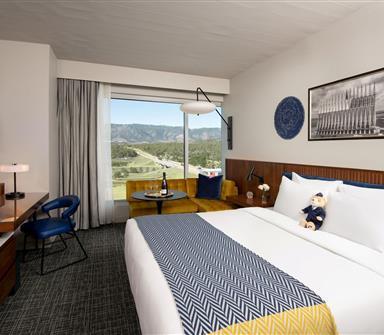 Mountain-view king room with white bedding, blue headboard, blue-and-yellow patterned throw, wood wall paneling, mustard window sofa, and desk area.