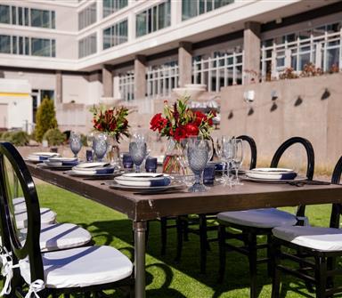 Outdoor table set for a meal with eight black chairs, white cushions, glassware, plates, and red floral centerpieces in front of a modern building.