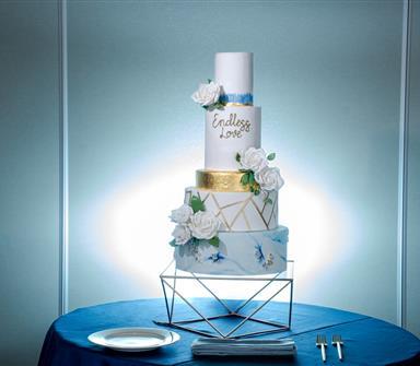 A four-tiered wedding cake with white roses and geometric gold accents sits on a blue tablecloth, with plates and forks beside it.