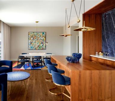 Contemporary suite living and dining area with large wood bar, blue velvet seating, brass pendant lights, colorful abstract artwork, and dark marble accent wall.