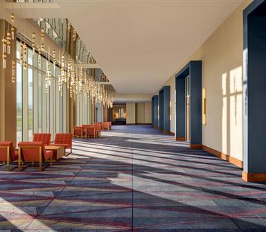 Sunlit hotel corridor with floor-to-ceiling windows, cascading pendant lights, coral lounge chairs, blue architectural accents, and patterned carpet.
